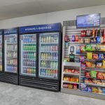 Three refrigerated drink coolers with various beverages are next to a snack display shelf stocked with chips, candy, and packaged snacks in a clean, modern room.