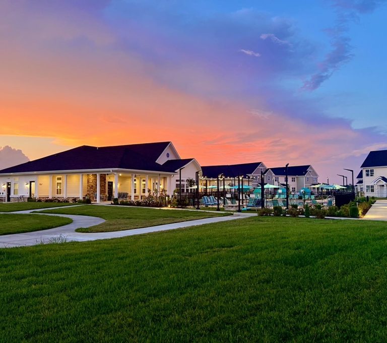 A residential complex with well-manicured lawns and multiple buildings at sunset, under a sky with orange and purple hues. It includes a pool area surrounded by a fence.