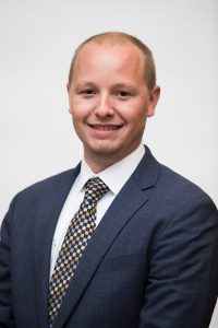 A man in a blue suit and patterned tie poses for a professional headshot against a plain white background.